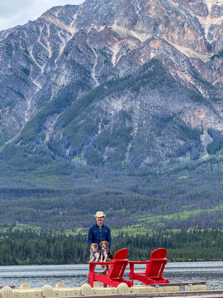 Man with two beagles on a floating dock beside red chairs, backed closely by a rocky mountain slope.