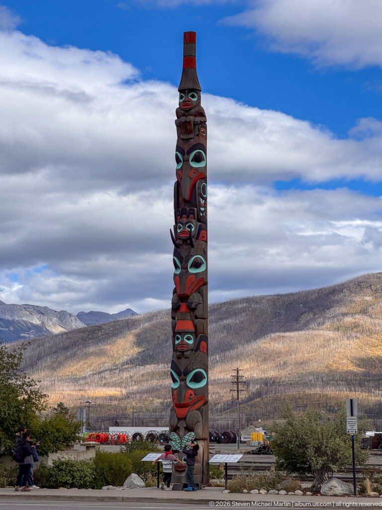 Tall carved and painted totem pole with red, black, and turquoise figures against mountains and a blue-and-cloudy sky.