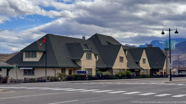 Steep-roofed gabled commercial building at a Jasper street corner with crosswalks, storefronts, clouds, and mountains.