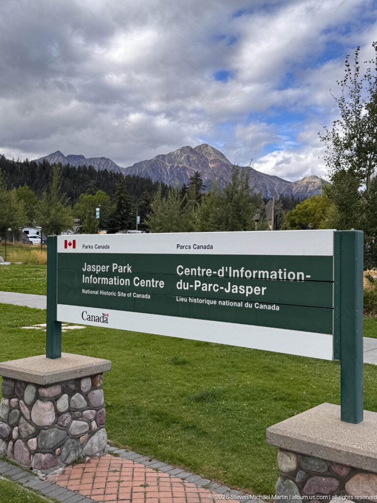 Bilingual Jasper Information Centre National Historic Site sign on a lawn with trees and a mountain ridge behind.