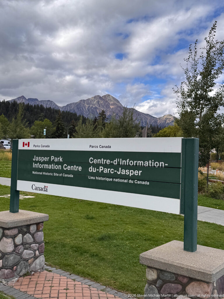 Green-and-white Parks Canada sign for Jasper Information Centre National Historic Site of Canada with mountains behind.