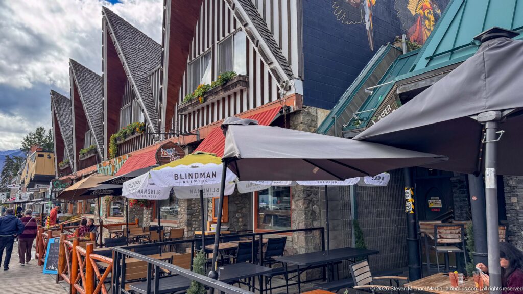 Downtown Jasper restaurant row with patio umbrellas, awnings, stone storefronts, steep gables, and pedestrians.