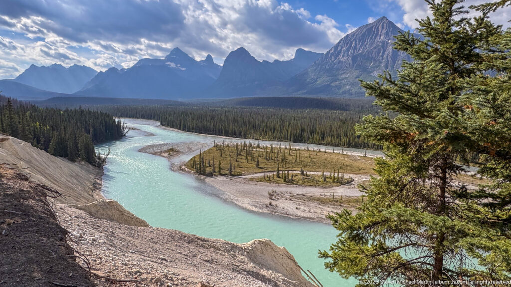 Turquoise river curving around gravel bars and conifer forest below a jagged Canadian Rockies mountain range.