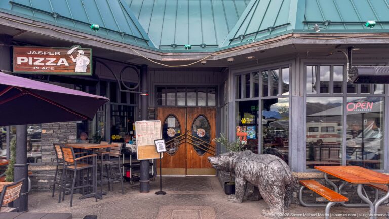 Jasper Pizza Place entrance with wooden double doors, open sign, patio tables, green roof, and a large bear sculpture.