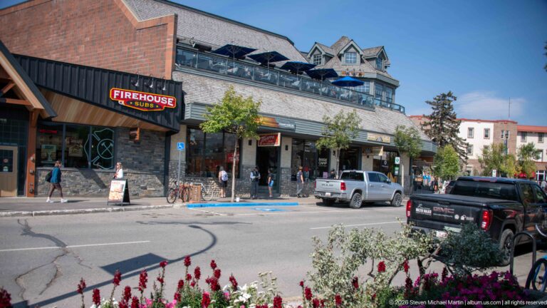 Downtown Jasper commercial block with Firehouse Subs sign, stone storefronts, upper terrace, parked vehicles, and foreground flowers.