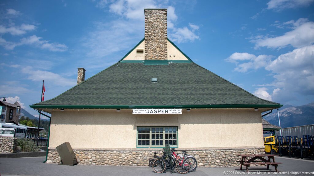 End elevation of Jasper railway station with green hipped roof, stone chimney, stucco wall, fieldstone base, bicycles, and mountains.