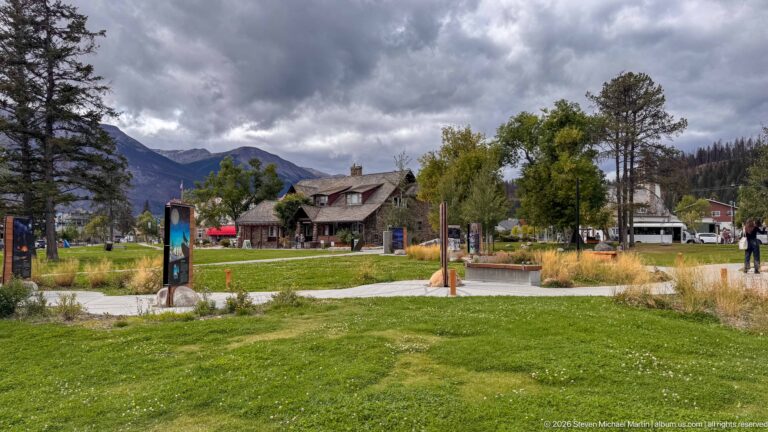 Landscaped lawn and curving paths leading toward the Jasper Information Centre beneath dark clouds and mountain slopes.