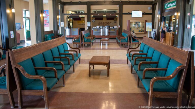 Facing rows of wooden benches with green upholstery and a small center table inside Jasper railway station.