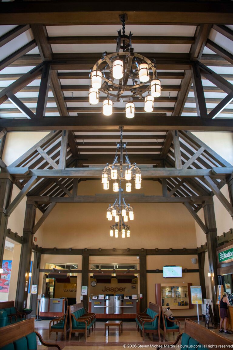 Upward view across layered timber trusses and hanging multi-light fixtures in Jasper railway station waiting room.