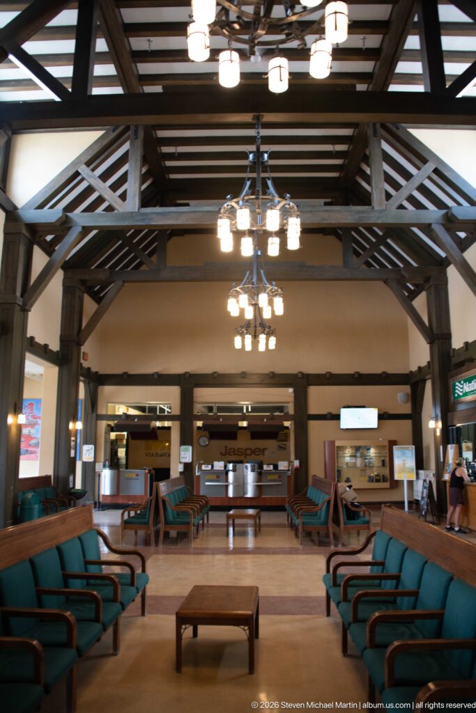 Wide symmetrical Jasper station waiting room with green benches, exposed timber trusses, hanging lights, and VIA Rail counters.