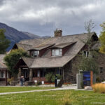 Three-quarter view of the Jasper Information Centre with stone walls, shingled roofs, dormers, lawn, visitors, and mountains.