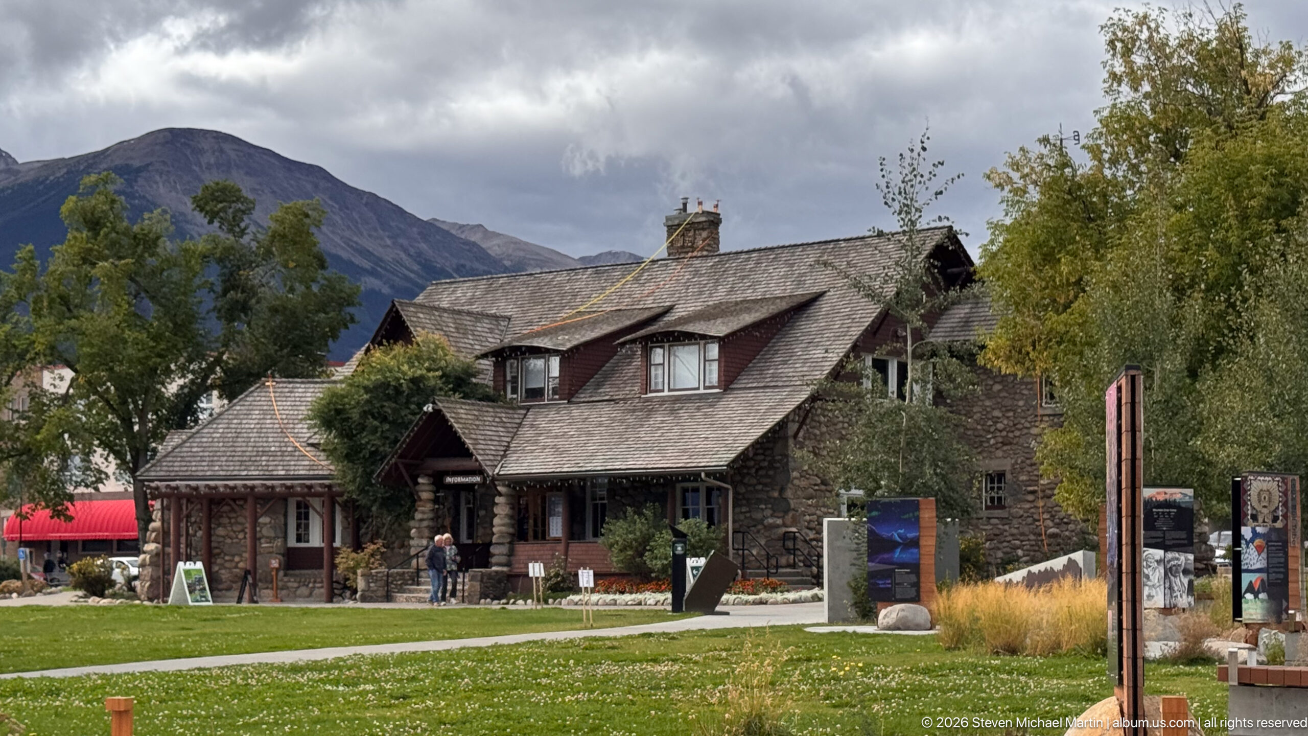 Three-quarter view of the Jasper Information Centre with stone walls, shingled roofs, dormers, lawn, visitors, and mountains.