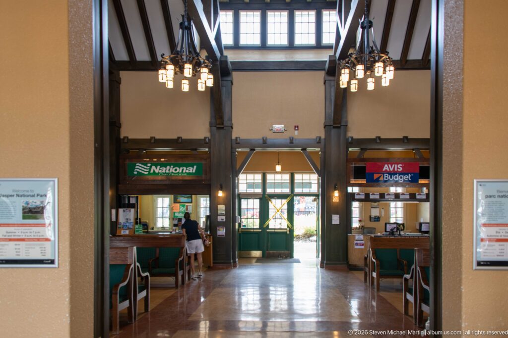 Centered view through Jasper station waiting room with timber posts, hanging lights, benches, rental counters, and green exit doors.