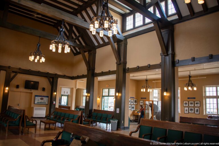 High waiting room with exposed timber posts and braces, hanging lights, clerestory windows, and green-upholstered wooden benches.