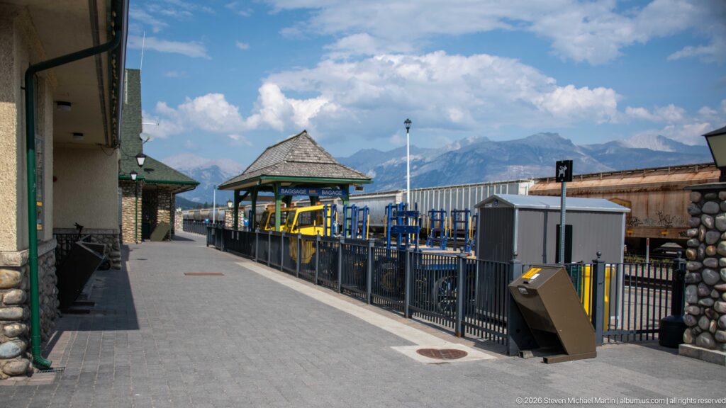 Jasper station platform with pavilion, railings and railway equipment, backed by mountains beneath a partly cloudy sky.