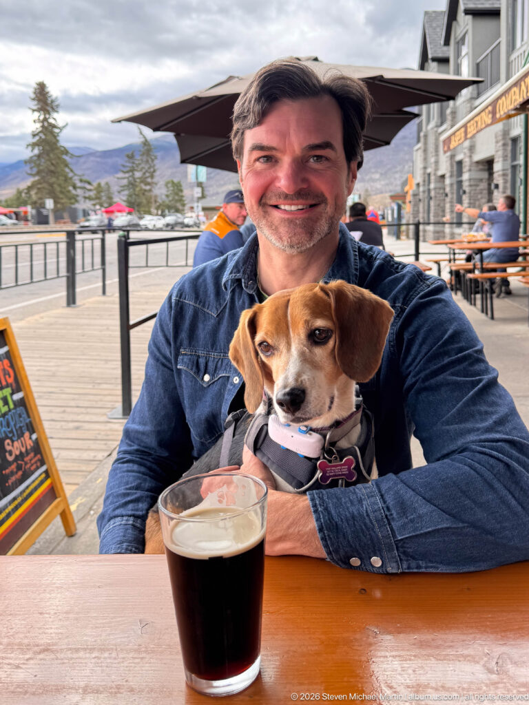 Man holding a tricolor beagle at an outdoor table with a glass of dark beverage, patio umbrellas, and Jasper storefronts behind.