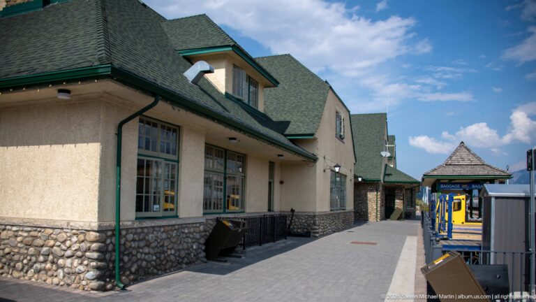 Platform-side exterior of Jasper railway station with fieldstone base, stucco walls, green trim, steep roofs, and a small pavilion.