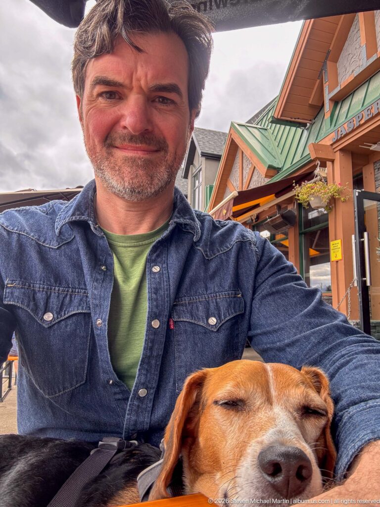 Seated man in a denim shirt with a tricolor beagle resting against him at an outdoor Jasper café table.