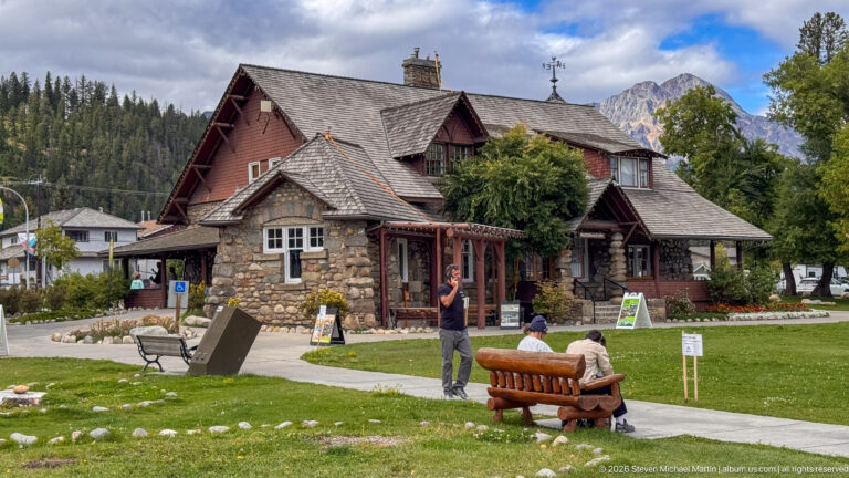 Jasper Information Centre with stone walls, shingled gables, timber details, lawn, benches, visitors, and mountains behind.
