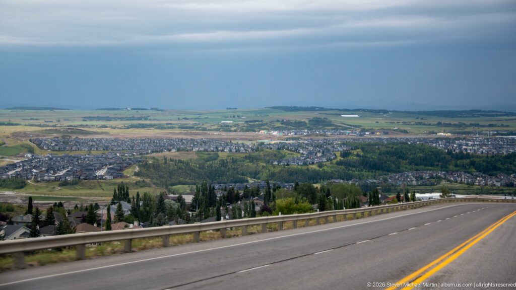 Elevated highway view over Rocky Mountain House neighborhoods, wooded areas, and farmland beneath a dark stormy sky.
