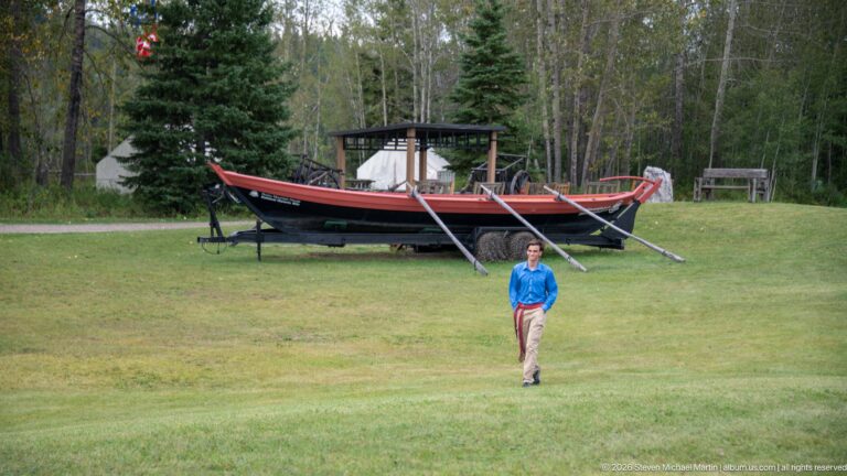 Man walking across grass with a large wooden boat display, tents, and trees behind him.