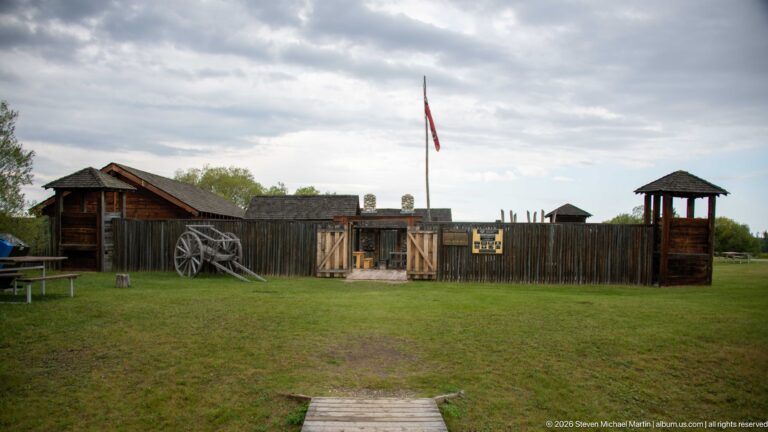 Frontal wide view of a timber fort enclosure across a lawn with central gate and flagstaff.