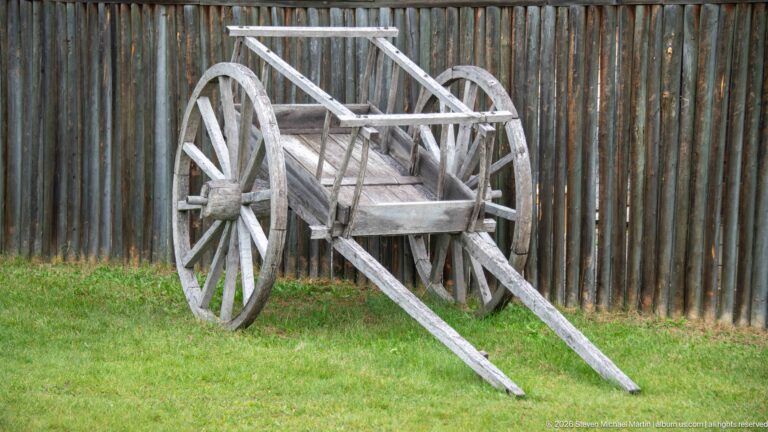 Weathered two-wheeled wooden cart with long shafts beside a vertical log palisade.