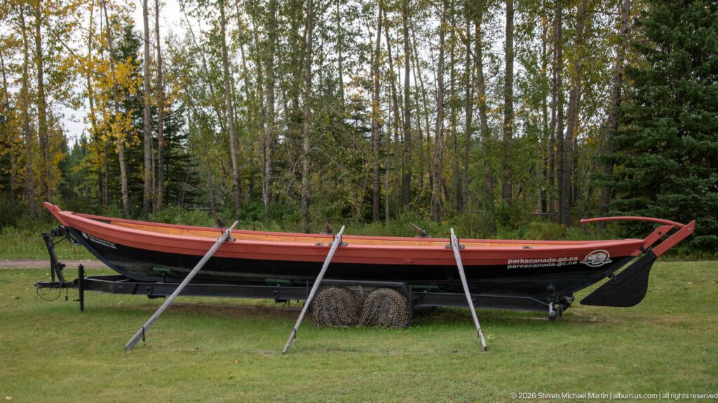 Long open wooden fur-trade boat replica on supports with several long oars beside it.