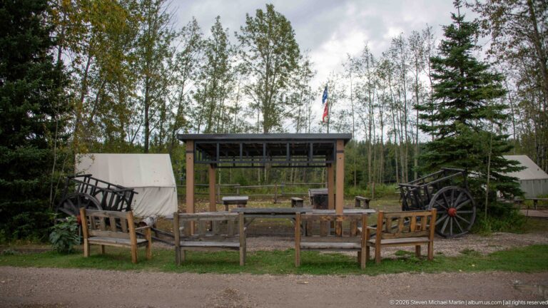 Canvas tents, wooden wagon, benches, and a roofed shelter in a wooded interpretive camp.