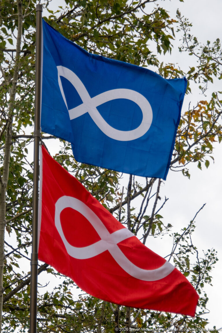 Blue and red Métis flags with white infinity symbols flying against leafy branches.