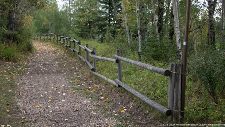 Gravel woodland trail curving beside a split-rail fence with fallen autumn leaves.