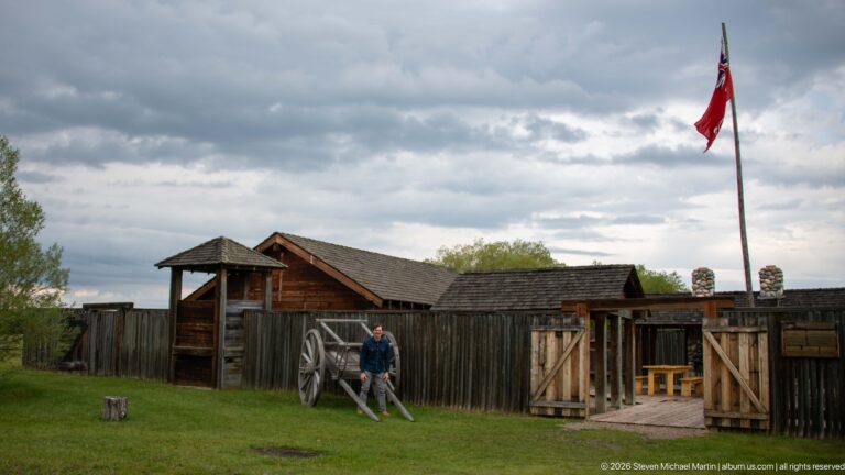 Wide view of a timber fort enclosure with open gate, visitor, flagstaff, and cloudy sky.