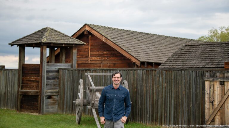 Man standing before a reconstructed timber palisade with shingled buildings and a wooden wheel.