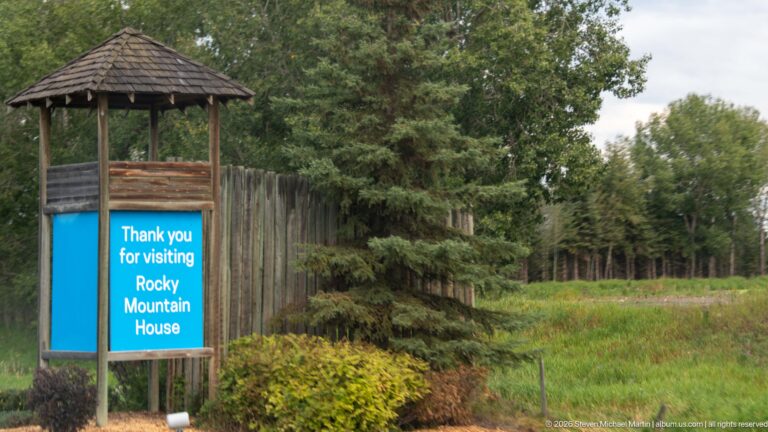 Blue Rocky Mountain House thank-you sign beside a tall log palisade and trees.