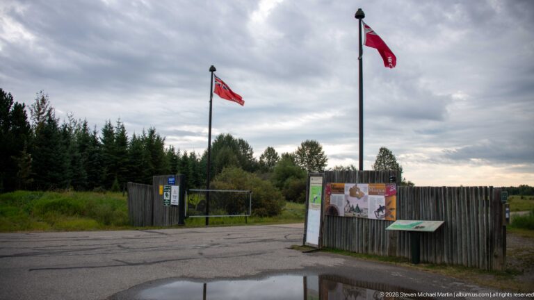 Entrance area with two Canadian flags, rustic vertical-log fencing, interpretive panels, gate, and puddle under cloudy sky.