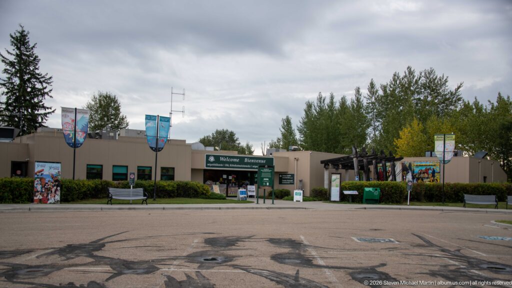 Wide view of low visitor-centre buildings with banners, murals, landscaping, entrance signs, and timber walkway.