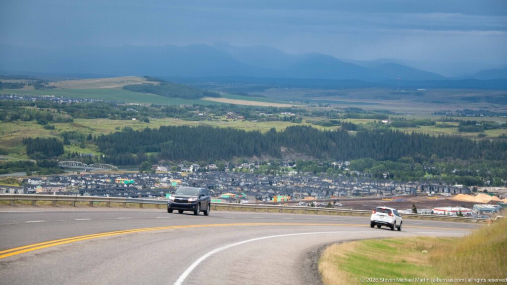 Highway curving downhill toward a town and green valley, with cars on the road and hazy mountains beneath dark clouds.