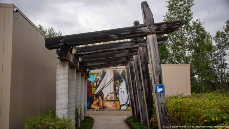 Rustic timber pergola over a paved walkway leading toward a colorful tipi mural beside the visitor building.