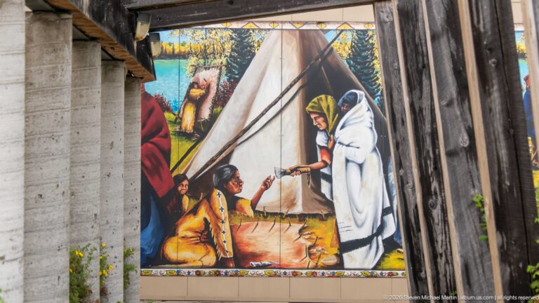 Close view of colorful tipi mural with Indigenous figures, partly framed by heavy weathered timber posts.