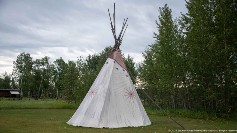 Full-height pale canvas tipi with painted starbursts and tall projecting poles standing on grass before a line of trees.