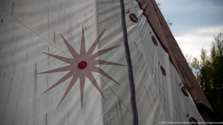 Close view of pale tipi canvas painted with a large brown starburst, red center, and smaller oval motifs beside a dark pole.