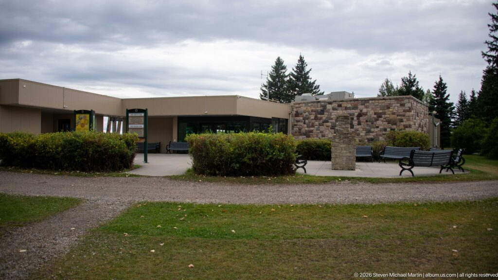 Low modern visitor centre behind hedges and gravel paths, with stone-faced wall, benches, and bicycle racks near the entrance.