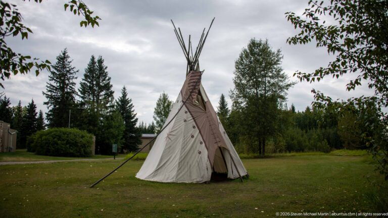 Painted canvas tipi with tall poles standing on an open lawn among trees beneath a cloudy sky.