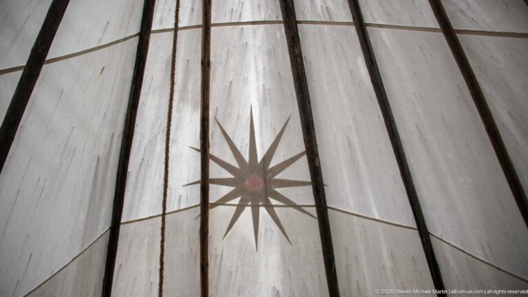 Large brown starburst with red center painted on pale tipi canvas between two dark vertical lodge poles.