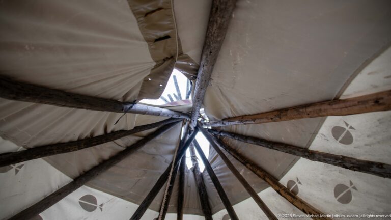 Upward interior view of dark wooden tipi poles converging at the smoke opening beneath pale canvas panels.