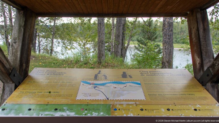Sloping interpretive map beneath a rustic timber shelter, with trees and the river visible through the open sides.