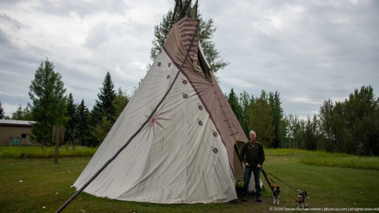 Side view of tall painted canvas tipi with projecting poles, with a man and two small dogs standing at its base.
