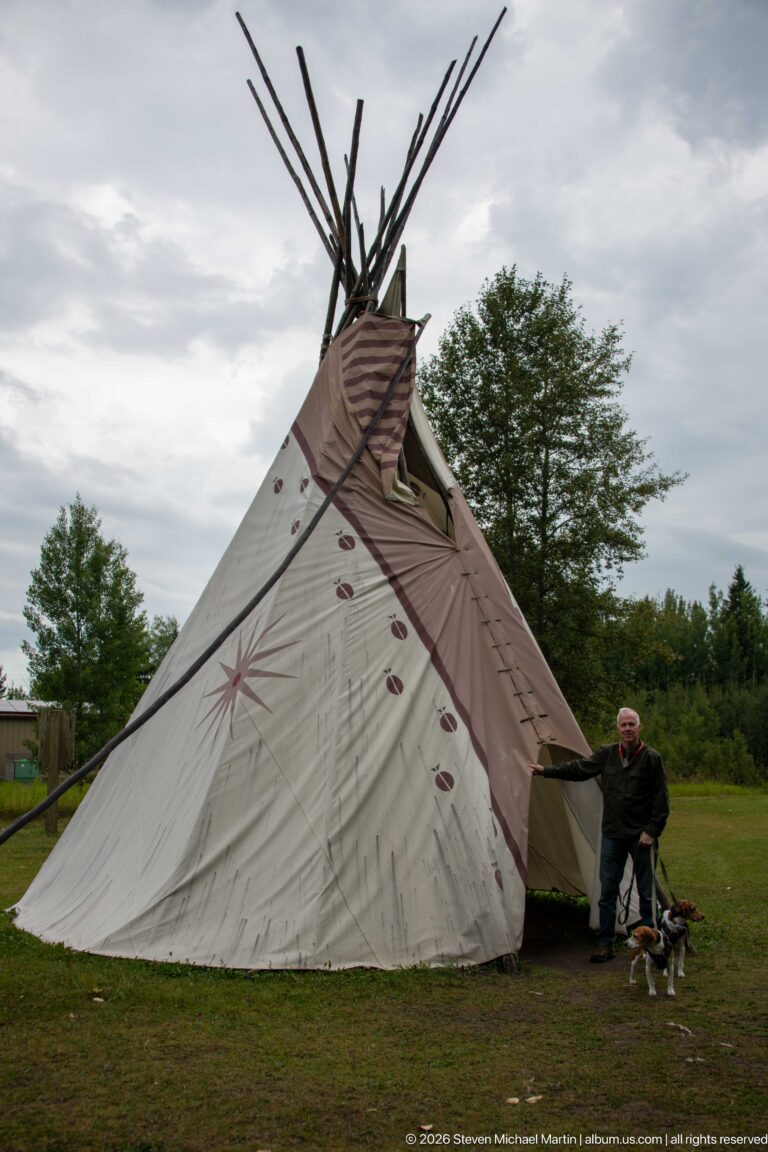 Large painted canvas tipi with tall lodge poles; a man and two small dogs stand beside its open entrance.
