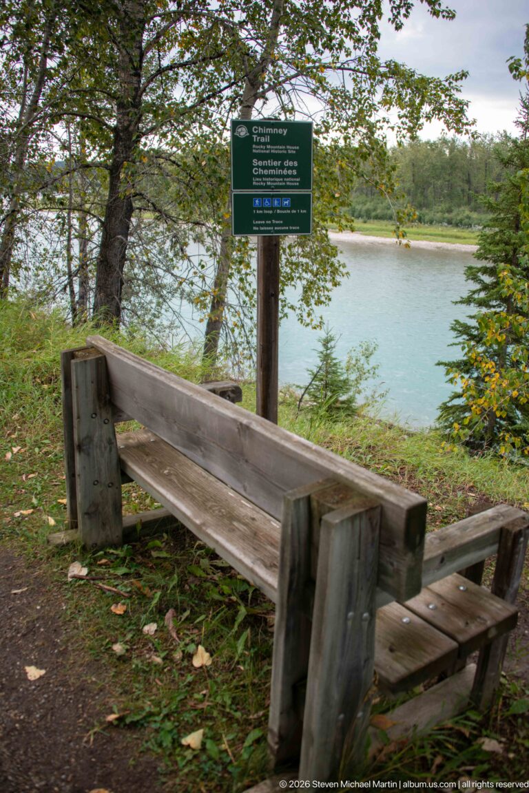 Rustic wooden bench beneath trees beside a green interpretive sign overlooking the blue-green North Saskatchewan River.