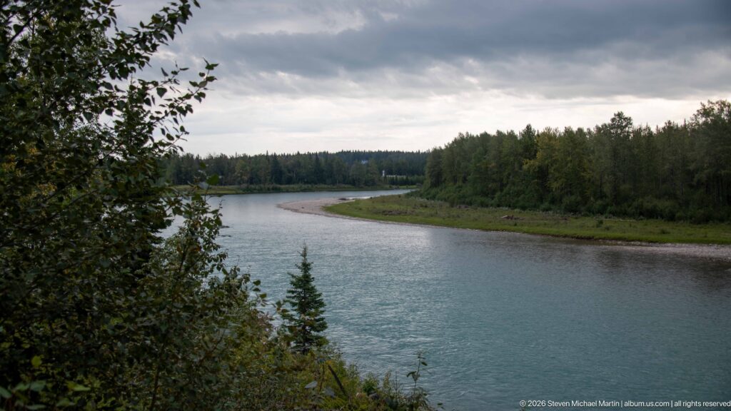 Broad blue-gray river bend with gravel bar and dense forest beneath a dark overcast sky.